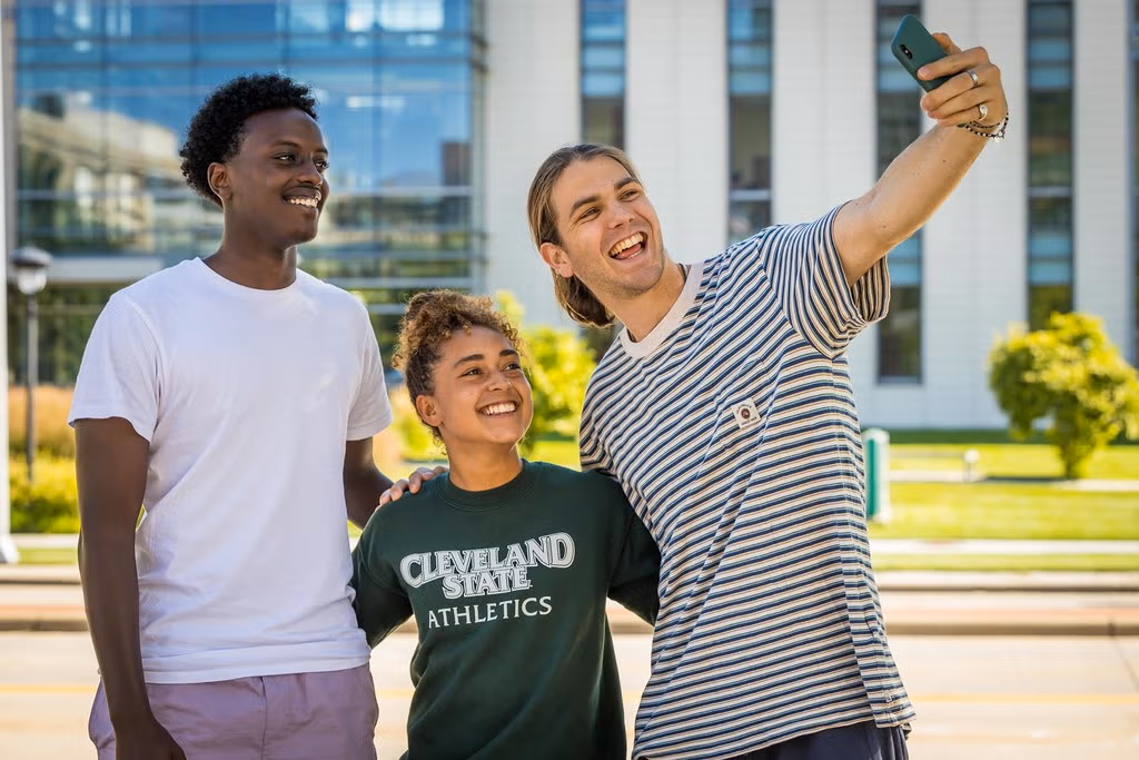 A group of three people stands outside a modern building while one of them takes a selfie. The person in the middle wears a Cleveland State Athletics shirt.